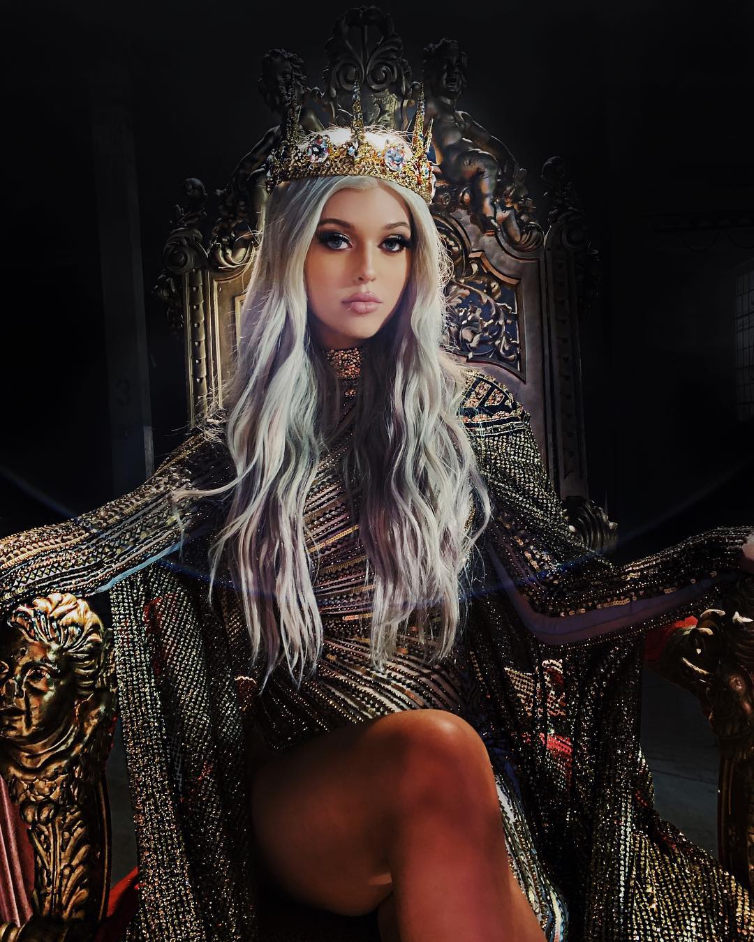 a woman with silver hair, adorned with a crown, sits on a golden throne. she wears a shimmering gold and black gown with elaborate jewelry, exuding an air of royal elegance and luxury. the setting is dark and dramatic, emphasizing her regal presence.