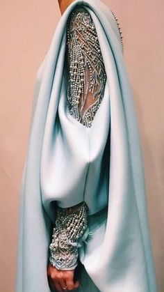 a woman is wearing a light blue gown with a silver beaded sleeve, which is beautifully patterned and elegant. the dress is flowing and voluminous, creating a dramatic and luxurious look. the model is posing with her hand resting on her hip, showcasing the intricate detail of the beaded sleeve. the image captures the essence of high fashion and glamor.