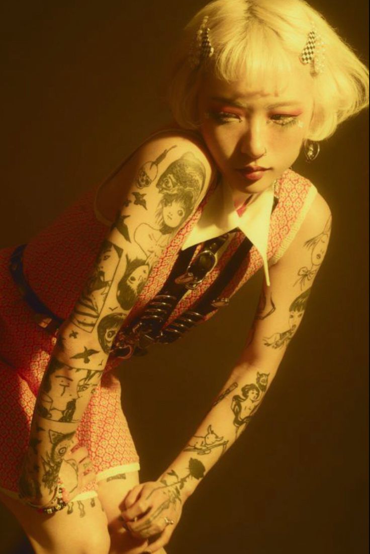 loseup shot of a young woman with short blonde hair and numerous tattoos on her arms. she's wearing a red and white patterned dress and various accessories. the lighting is warm and sepiatoned, creating a moody and artistic atmosphere. the woman's expression is pensive, and her gaze is directed downwards. the overall aesthetic is alternative and edgy.