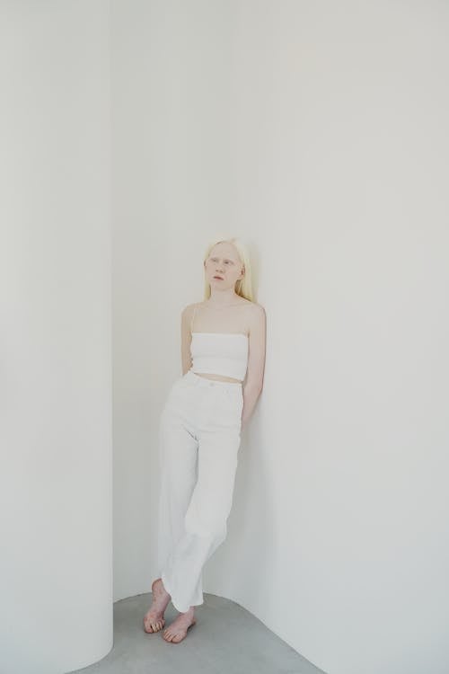 the image shows a young albino woman with blonde hair standing in a corner against a plain white background. she is wearing a white crop top and matching wideleg pants, creating a minimalist and monochromatic look. her pose is relaxed, yet her expression is serious and slightly pensive. the overall aesthetic is clean, modern and emphasizes the simplicity of the clothing and the model's striking features. the lighting is soft and even, highlighting the texture of the clothes and the model's skin.