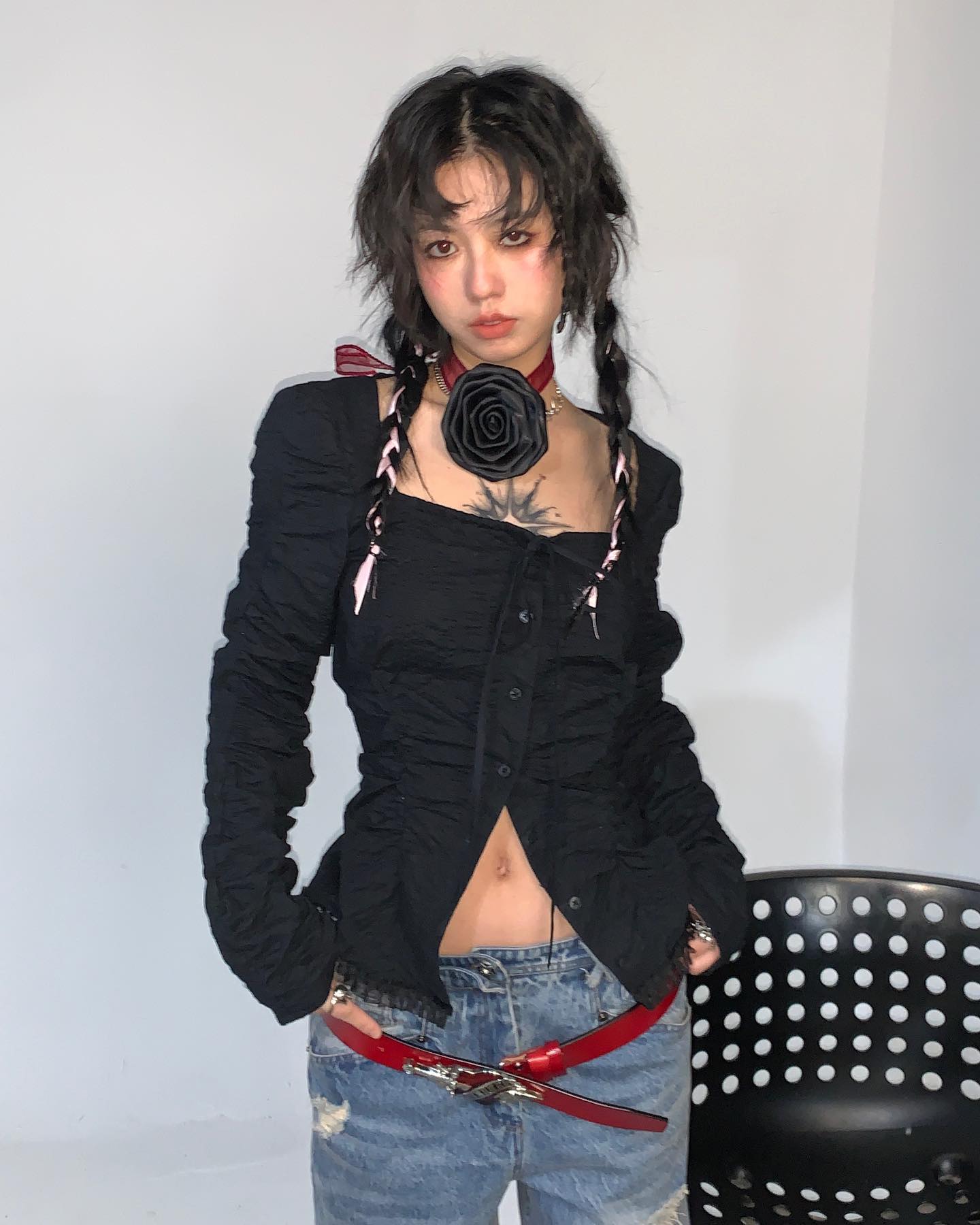 a young woman with dark hair styled in braids is posing in a studio shot. she's wearing a black, ruched, longsleeved shirt with a square neck, and highwaisted jeans.  she has a red belt and a black rose necklace. her style is edgy and alternative, with a hint of gothic and grunge aesthetics.  the overall image is stylish and welllit.