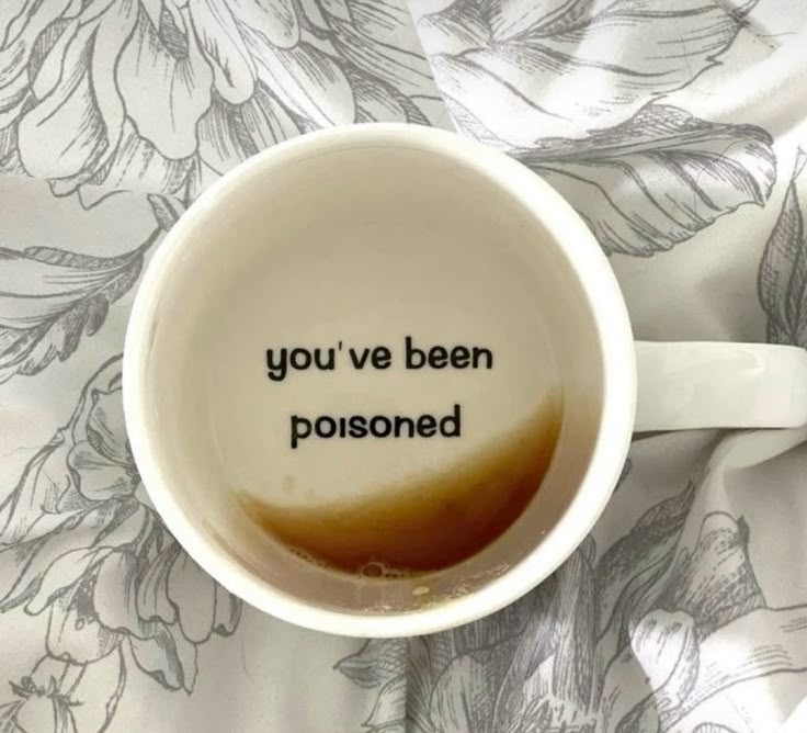 a mug with the words "you've been poisoned" printed on the inside, with a small amount of coffee or tea remaining. the mug sits on a floralpatterned bed sheet or tablecloth.