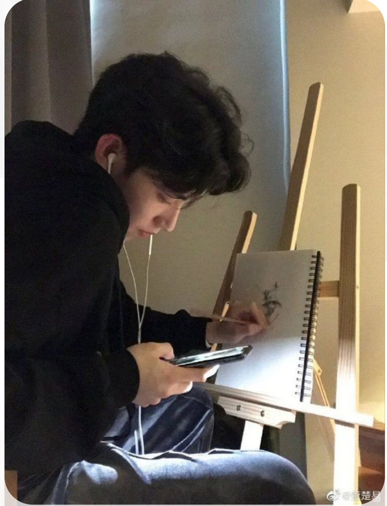 a young man with dark hair is sitting in a chair, drawing on a sketchpad propped on a wooden easel. he's wearing a black hoodie and black pants, with white earphones in his ears. he's also holding a smartphone in his left hand. the background is a beige wall, and the room is lit by soft, warm indoor lighting.
