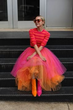 a young woman wearing a pink and orange tutu skirt and a pink and white striped sweater sits on the steps outside. she is wearing sunglasses and looks toward the left side of the frame.