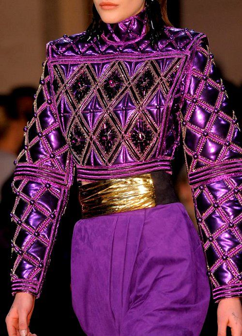 closeup view of a woman's upper body wearing a stunning purple metallic jacket. the jacket is richly embellished with beads and features a quilted diamond pattern. a wide gold belt cinches the waist, and the overall style is luxurious and elegant. the image appears to be from a fashion runway or photoshoot. the model's face is partially visible, but the focus is mainly on the intricate detail of the jacket. the color palette is dominated by deep purple and gold.