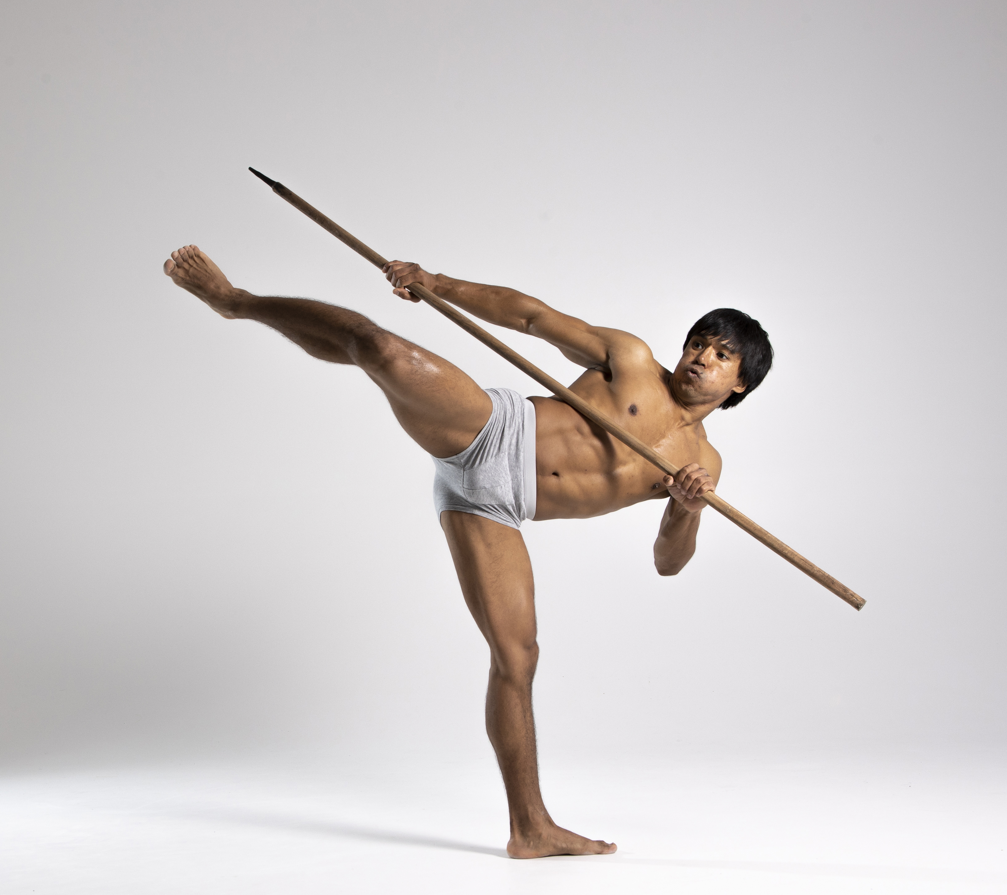 a barechested man wearing underwear stands in a studio setting, holding a spear while performing a kick. he is barefoot and balanced on one leg, with the other leg extended midair.