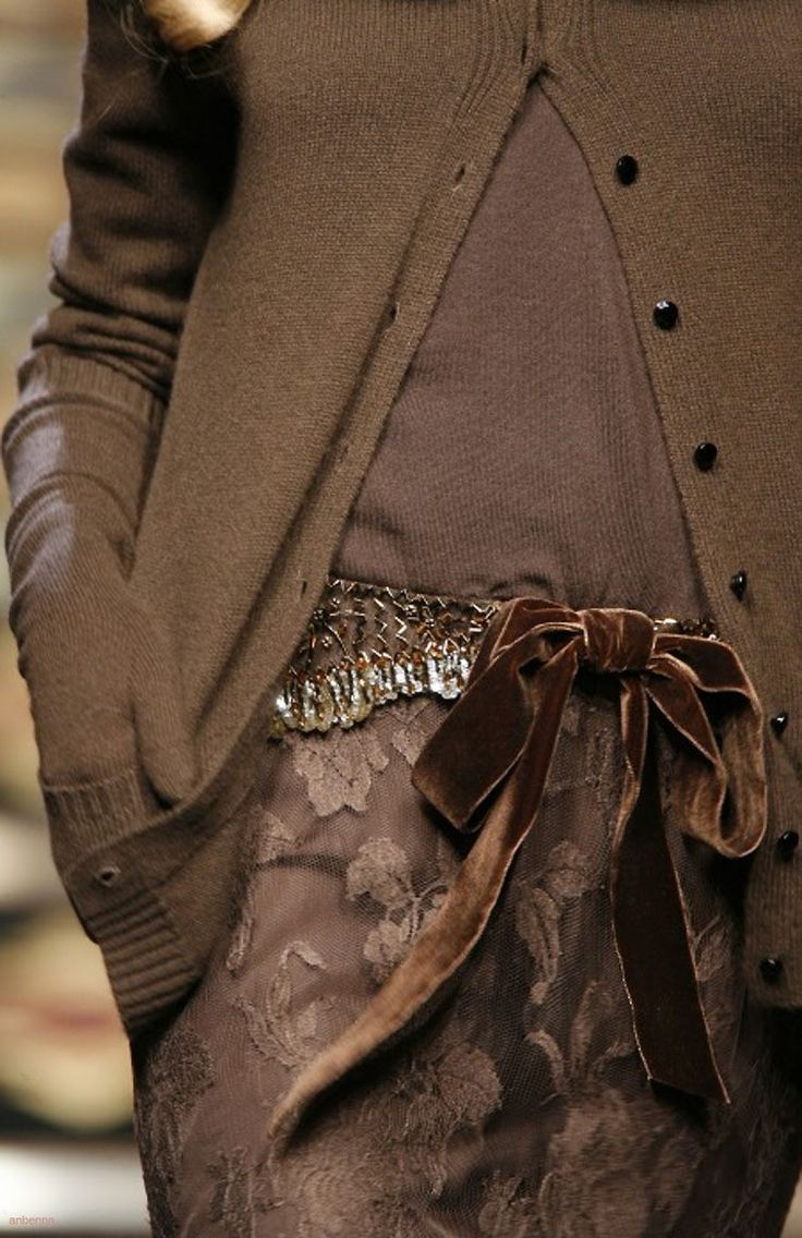 a closeup of a woman wearing a brown cardigan and a delicate brown lace dress with a matching velvet ribbon belt. the woman is standing with her arms crossed, showcasing the details of her stylish outfit. the ribbon is tied in a large bow and the overall look is elegant and sophisticated.