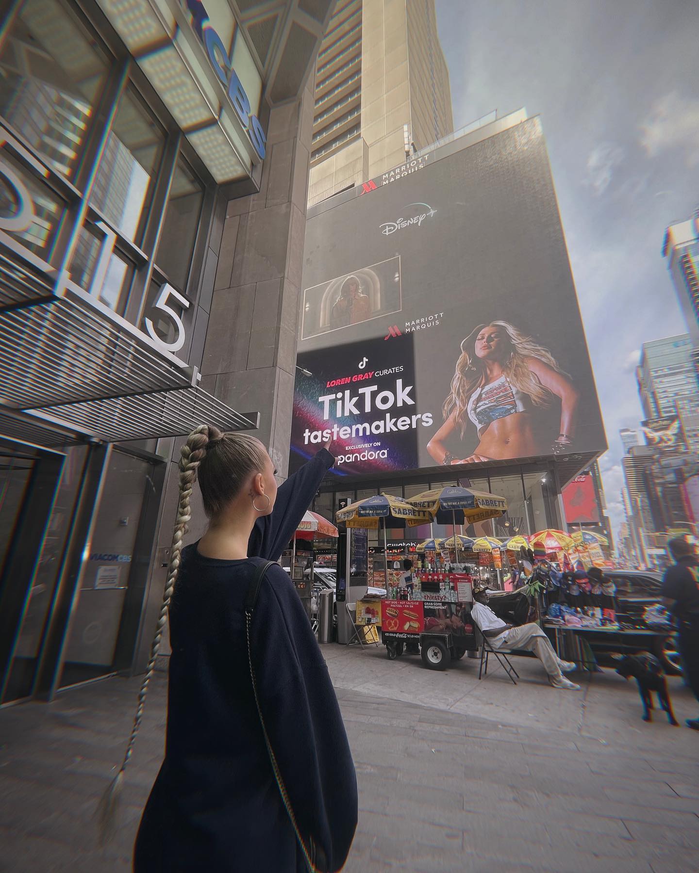a woman with a long braid is standing on a street in times square, pointing at a large billboard. the billboard features advertisements for disney+, tiktok, and pandora, and is located on the side of a building that appears to be the marriott marquis hotel. there are food carts and other people in the background, indicating a busy urban environment.