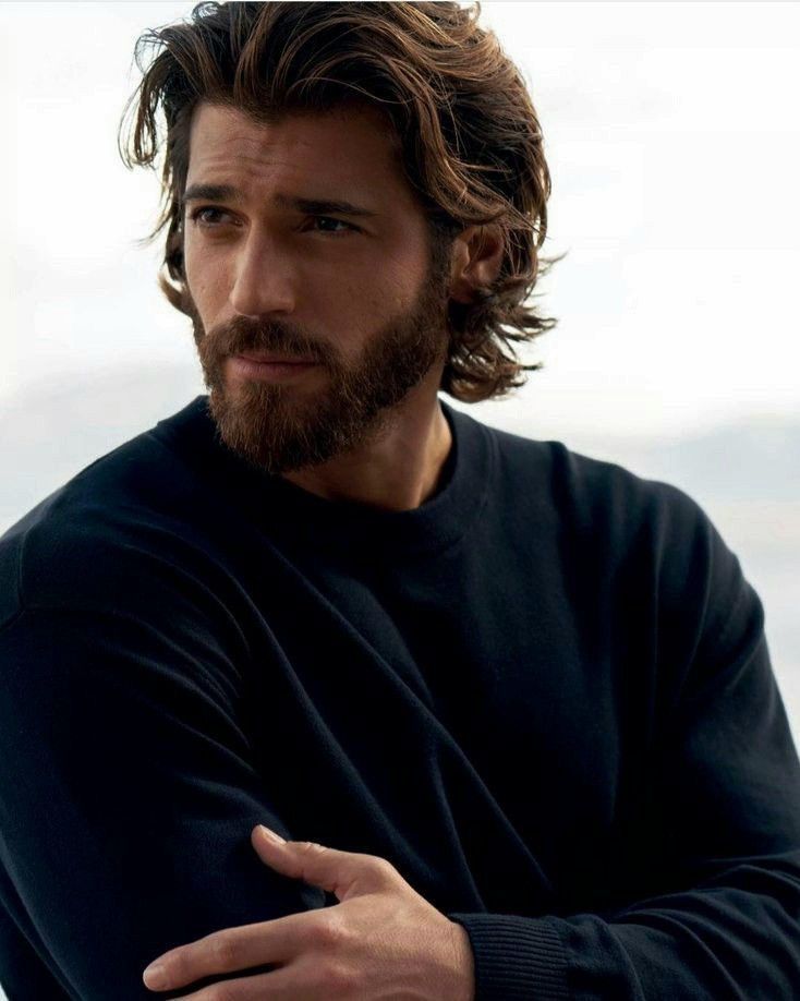 a portrait of actor and model can yaman. he has long wavy hair and a beard. he is wearing a black sweater and is looking off to the side.