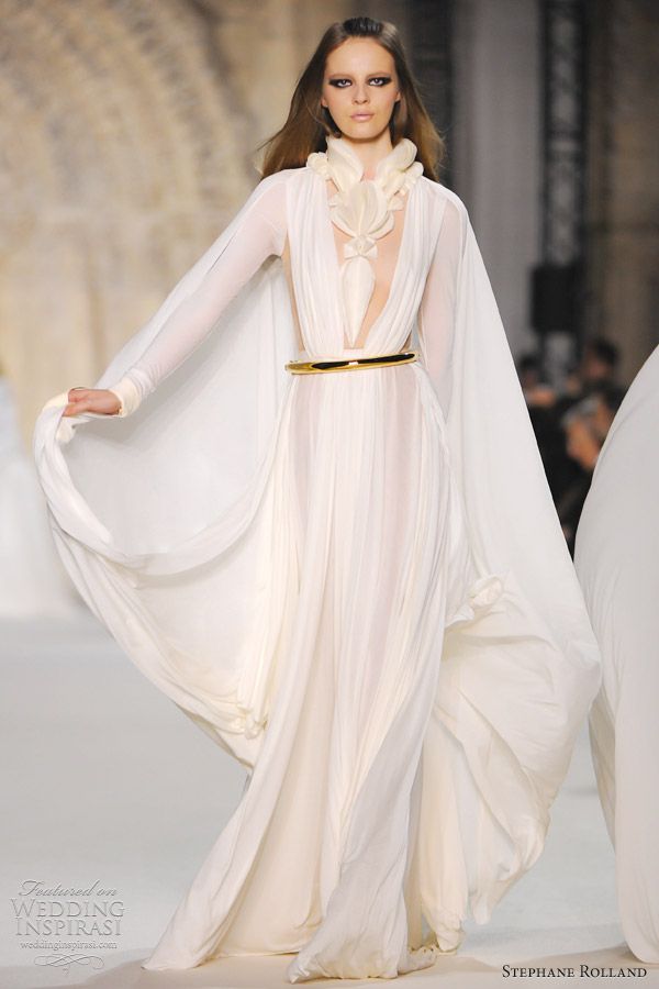 a woman in a white, flowing wedding dress is walking down a runway. the dress features a plunging neckline, a cape, and a gold belt. the model is wearing her hair down and has a natural makeup look. the dress is very ethereal and romantic, and the model looks stunning.