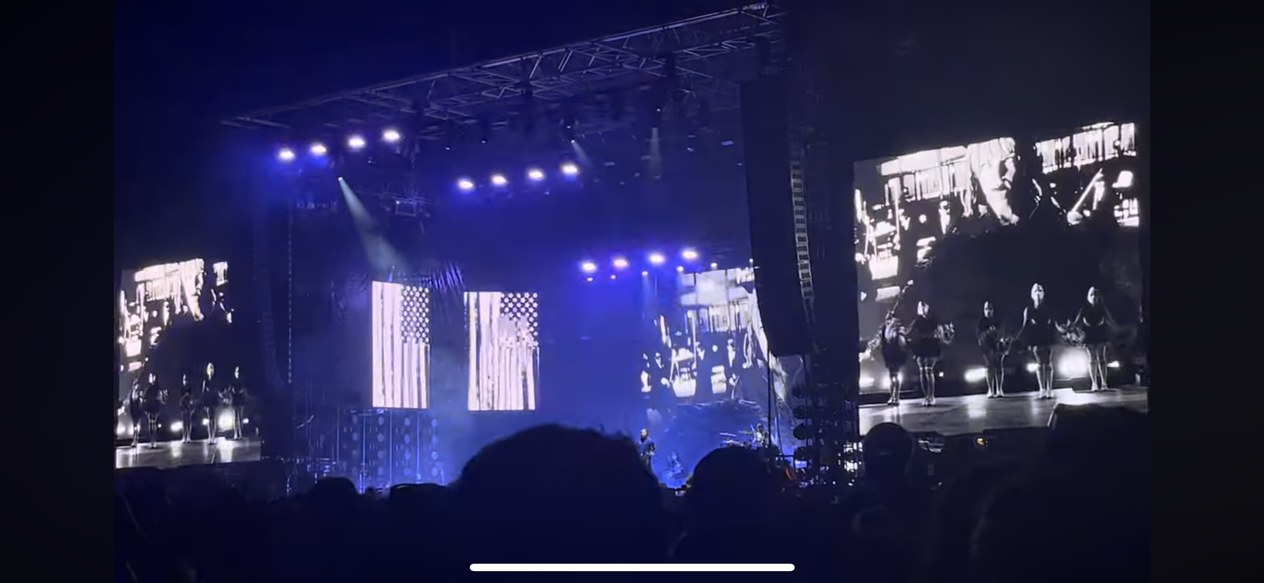 this is a concert shot from the audience of a band playing on stage in a dark venue. the band is playing in front of a giant screen with a blurry image of a band playing on stage and a black and white image of what appears to be an american flag. there are many bright lights shining on the stage, and the audience is excited and clapping their hands.