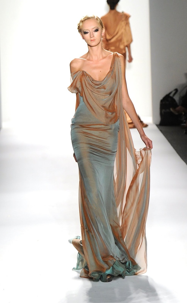 a fashion model walks down a runway wearing a flowing, sheer gown with ombre coloring in shades of teal and bronze. the dress is floorlength and drapes beautifully around her body.  the model has her hair pulled back and simple makeup.  another model in a similar toned dress is visible in the background, out of focus. the setting is a bright, minimalist runway.