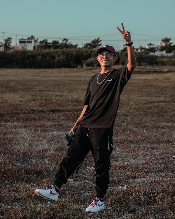 a young asian man stands in a field at sunset, smiling and giving a peace sign. he's wearing a black tshirt, black cargo pants, a maroon cap, glasses, and nike air force 1 sneakers.  the overall feeling is relaxed and casual.