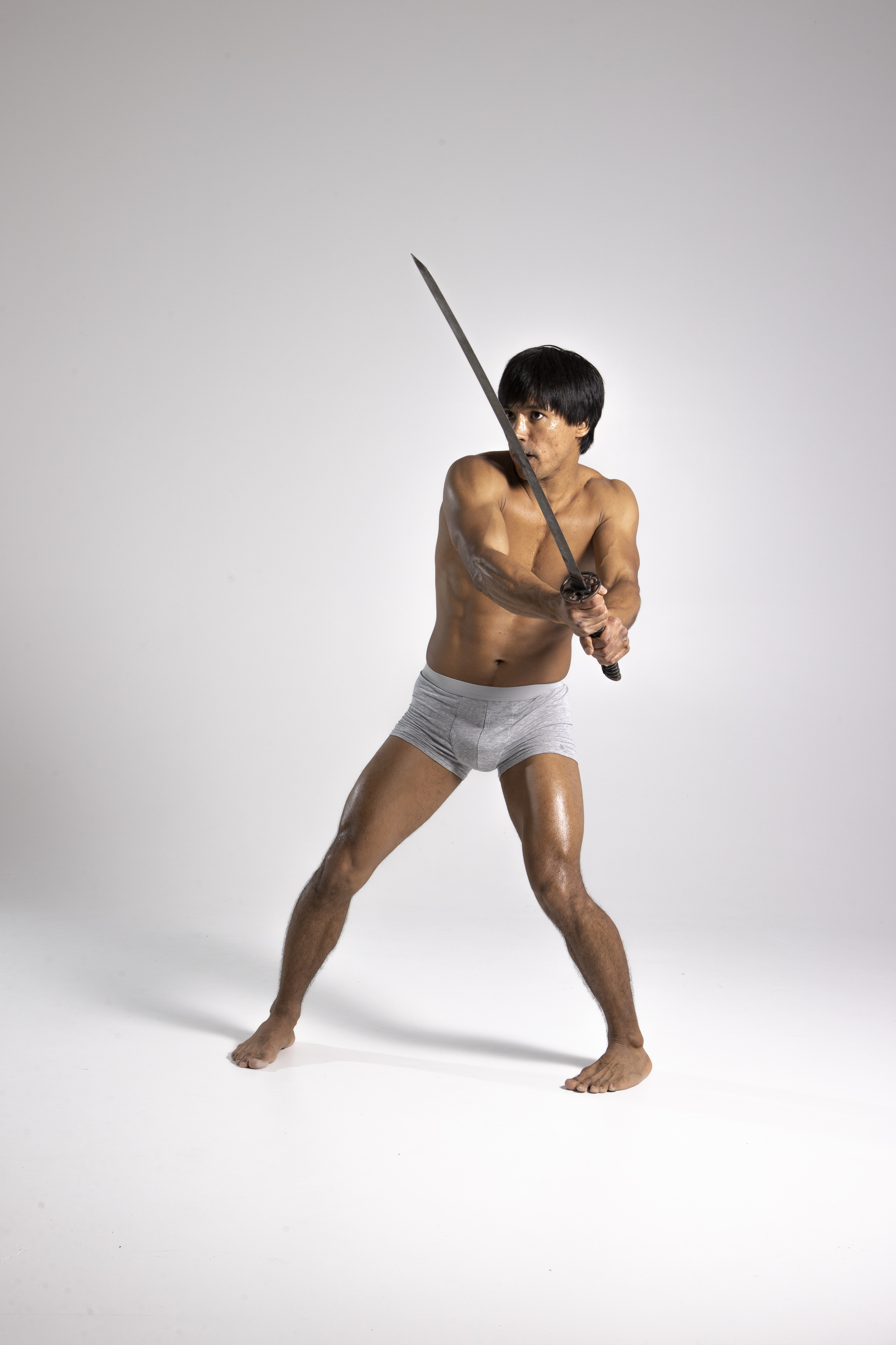 a shirtless man in grey underwear is holding a sword (katana) in a defensive pose. he has an asian appearance. he is barefoot and appears to be practicing martial arts. the background is a plain white studio.