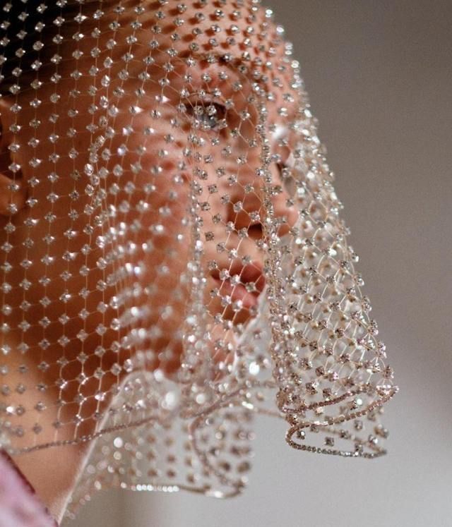 the image is a closeup shot of a model wearing a luxurious, sheer veil adorned with numerous small, sparkling rhinestones or crystals. the veil is intricately beaded, creating a delicate, almost ethereal effect. the veil covers the model's face partially, obscuring most of her features except for one eye and part of her mouth. the overall aesthetic is glamorous and elegant, suggestive of bridal wear or highfashion photography.  the background is blurred and out of focus, drawing attention to the detail and texture of the veil itself. the lighting is soft and diffused, enhancing the sparkle of the embellishments.