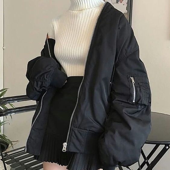 a woman is wearing a white turtleneck sweater, a black pleated skirt, and a black bomber jacket. the jacket is open and revealing the sweater underneath. the outfit is casual and fashionable.