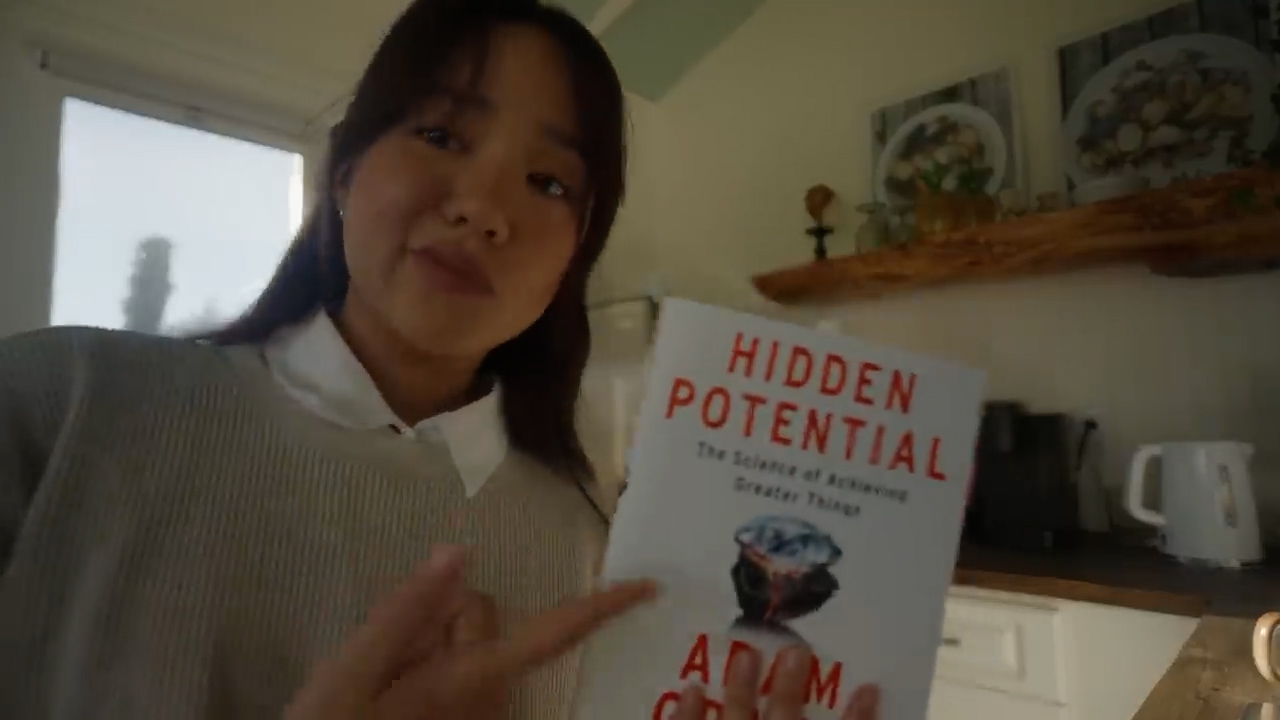 a young woman is holding up a copy of the book "hidden potential" by adam grant. she is indoors in a kitchen, and the lighting is natural and coming from a window behind her. the perspective is first person, as if the viewer is the one being shown the book.