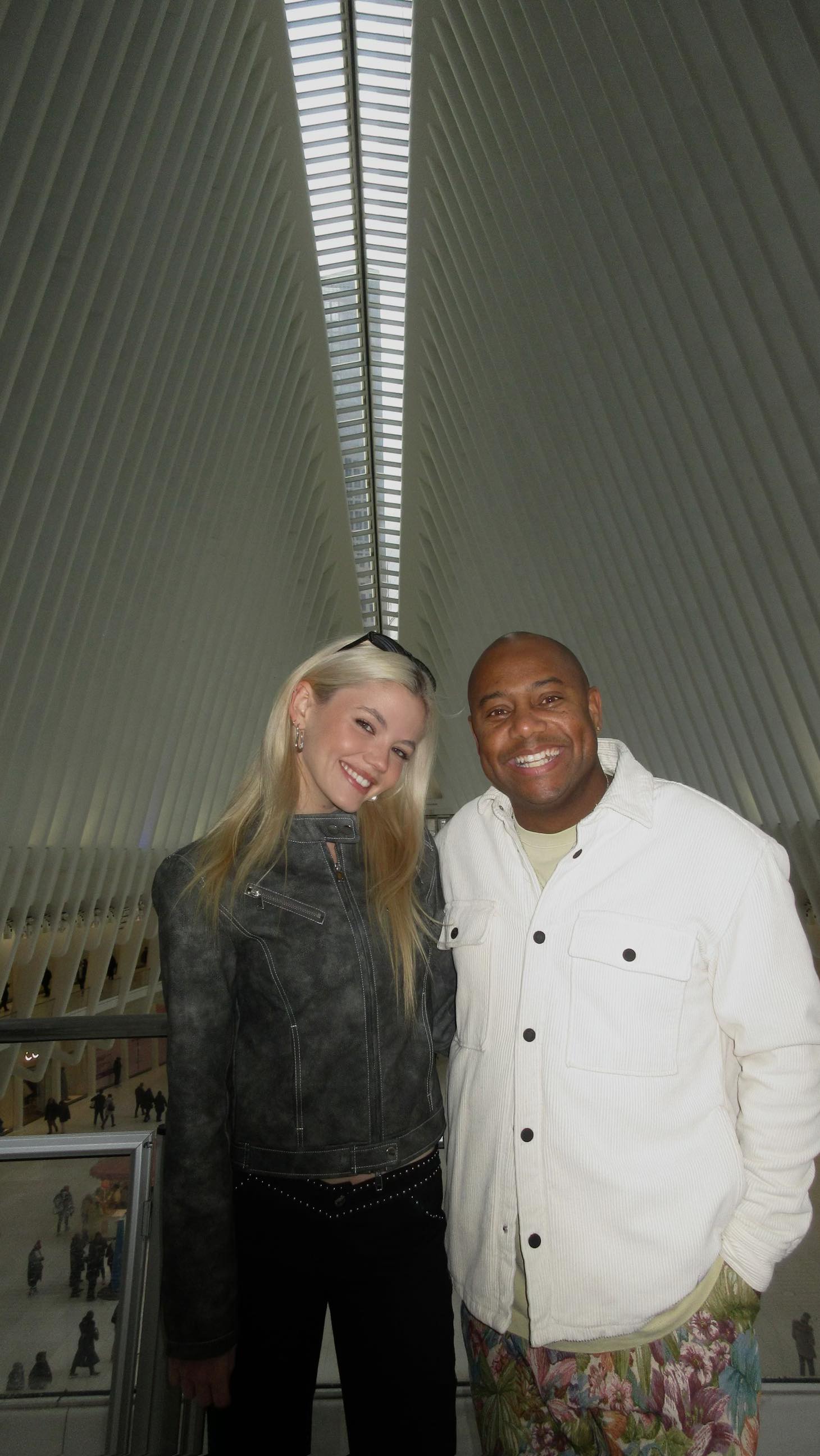 a photo of two people, a man and a woman, standing indoors with a unique ceiling design behind them. the woman has blonde hair, is wearing a gray jacket and black pants, and is smiling. the man is wearing a white shirt over a tshirt and floral pants, also smiling.