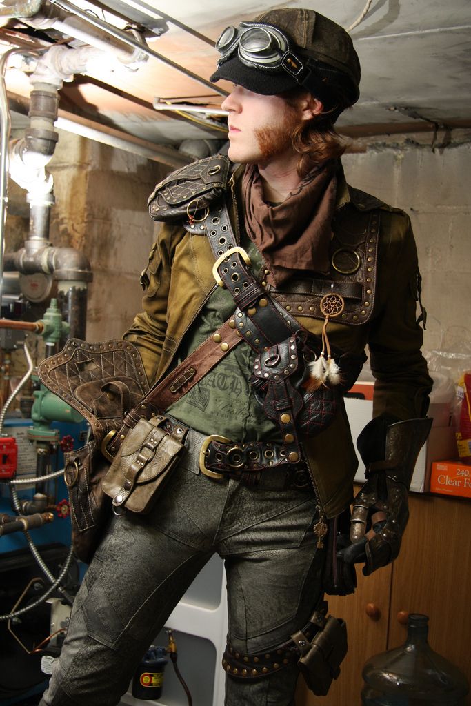a person dressed in a steampunk costume in an industrial setting. they are wearing a leather jacket, goggles, a belt with straps and metal accents, and pants.