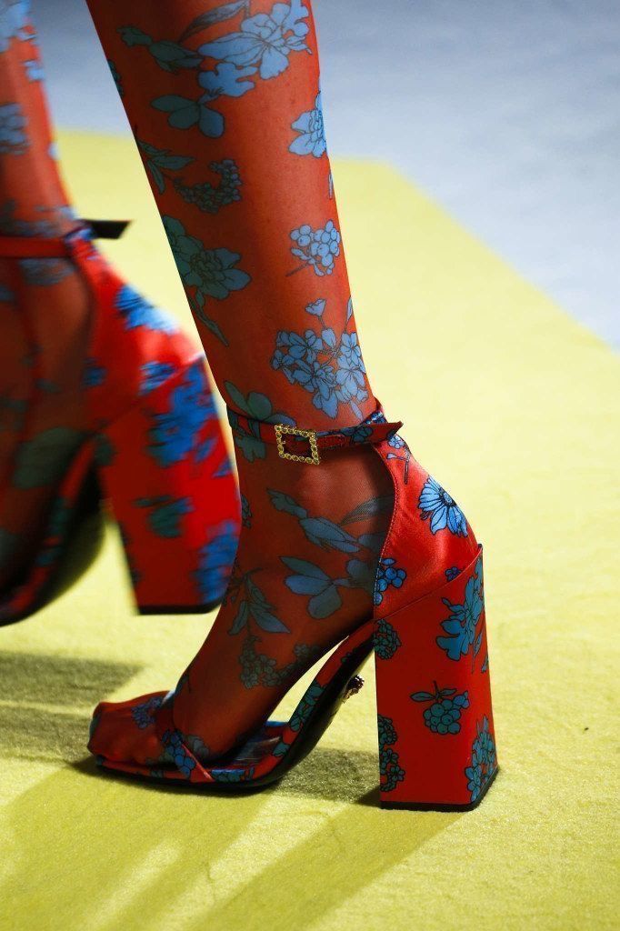 closeup of a woman's legs wearing floral printed red tights and red block heels with a gold buckle. the model is on a runway wearing the footwear.