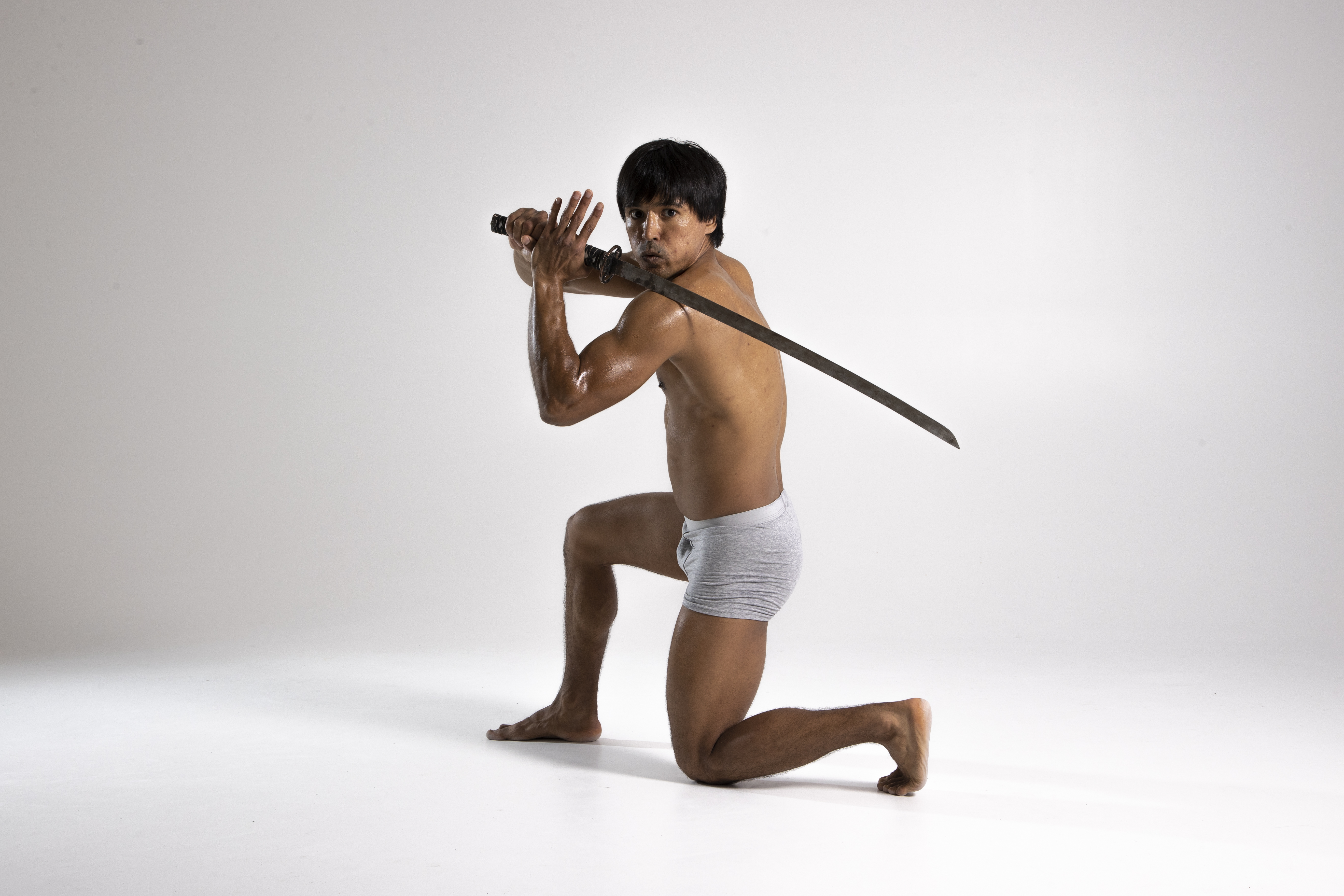 a barechested asian man is kneeling with one knee on the ground while holding a sword horizontally over his shoulder in a defensive posture. he is wearing grey underwear. the image is a studio shot with a white background. the man appears muscular and is posed in a martial arts or combat stance, holding the blade as if ready to defend himself.