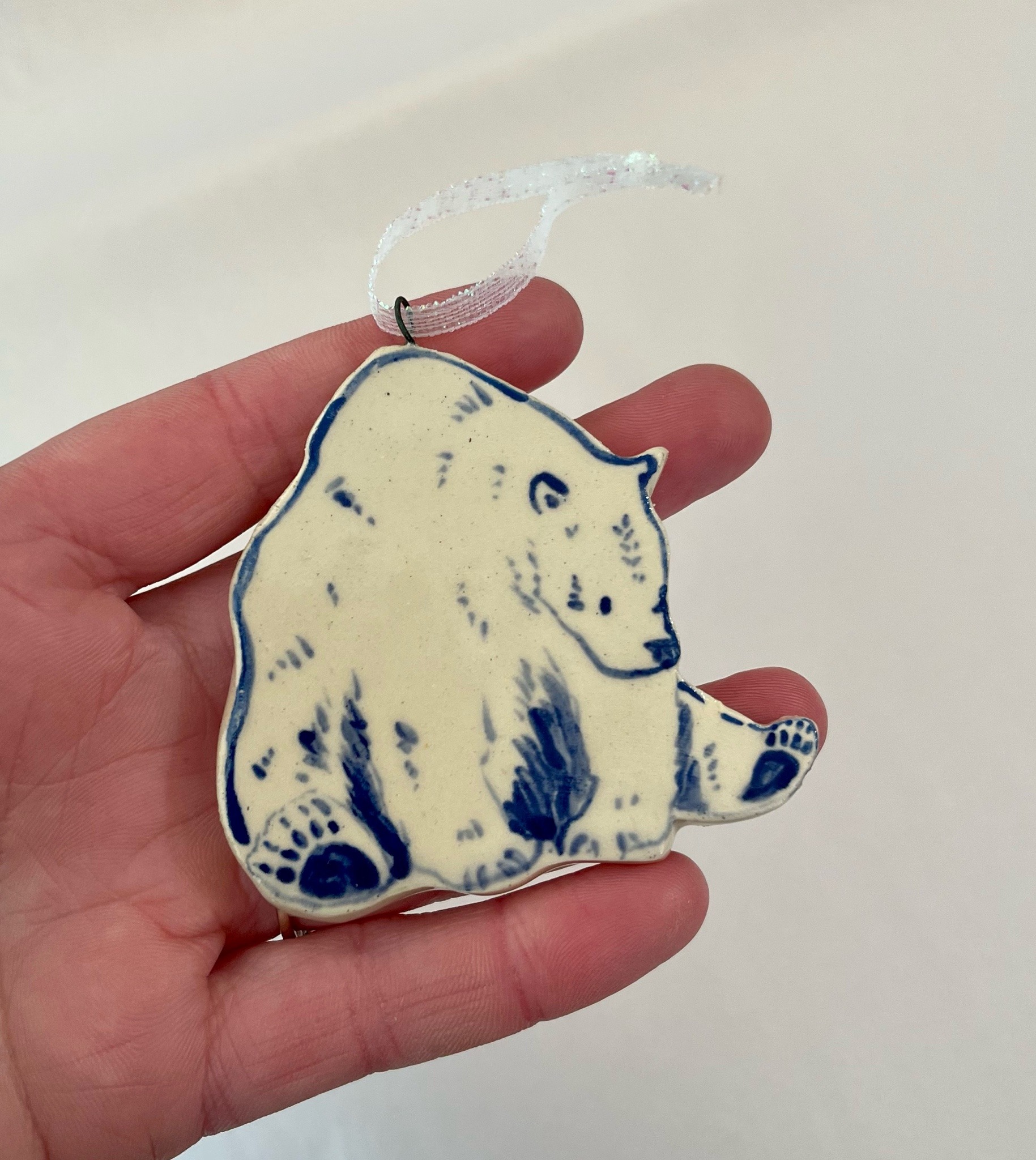 a closeup shot shows a hand holding a small, handmade ceramic ornament. the ornament is shaped like a polar bear sitting down,  painted in shades of white and blue. the bear's details are delicately handpainted.  the ornament has a simple wire loop at the top, attached to a white ribbon, suggesting it is designed to hang on a christmas tree or other display.  the background is a plain, offwhite surface. the hand holding the ornament provides a sense of scale.