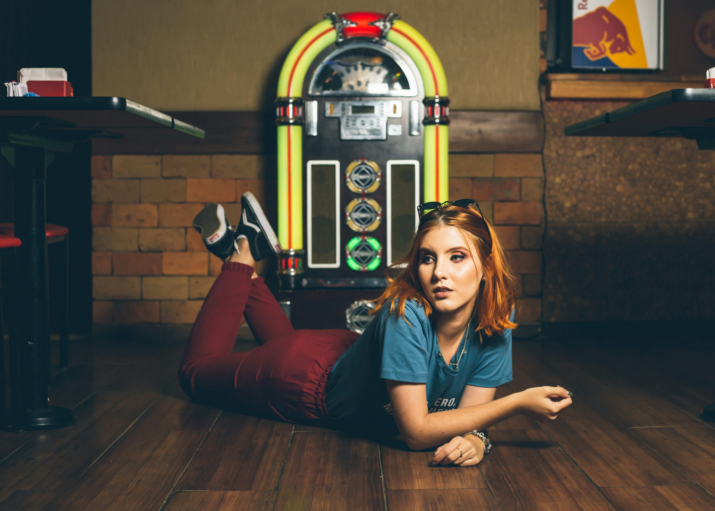 a young redhead woman is lying on a wooden floor in a retro setting. she is wearing red pants, a blue shirt, and sneakers. a vintage jukebox is visible in the background. the setting appears to be a bar or diner. the overall mood is relaxed and cool.