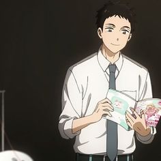 the image shows a young man with short dark hair and brown eyes, wearing a white shirt and a dark greenstriped tie. he is standing indoors, possibly in a classroom, and holding an open book with a cute, cartoonish design on the cover. the lighting suggests an indoor setting.