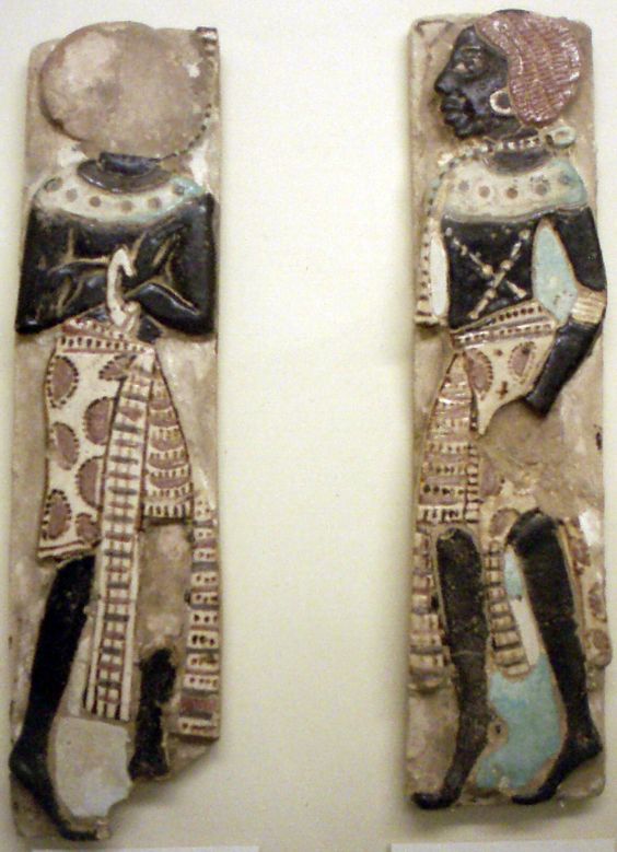 two ceramic fragments of figures, likely nubian, from ancient egypt. they are standing upright and facing forward, both with a raised hand. their bodies are mostly black with white and red accents. they both wear white and red patterned cloth, necklaces, and earings. the fragment on the left is missing its head.