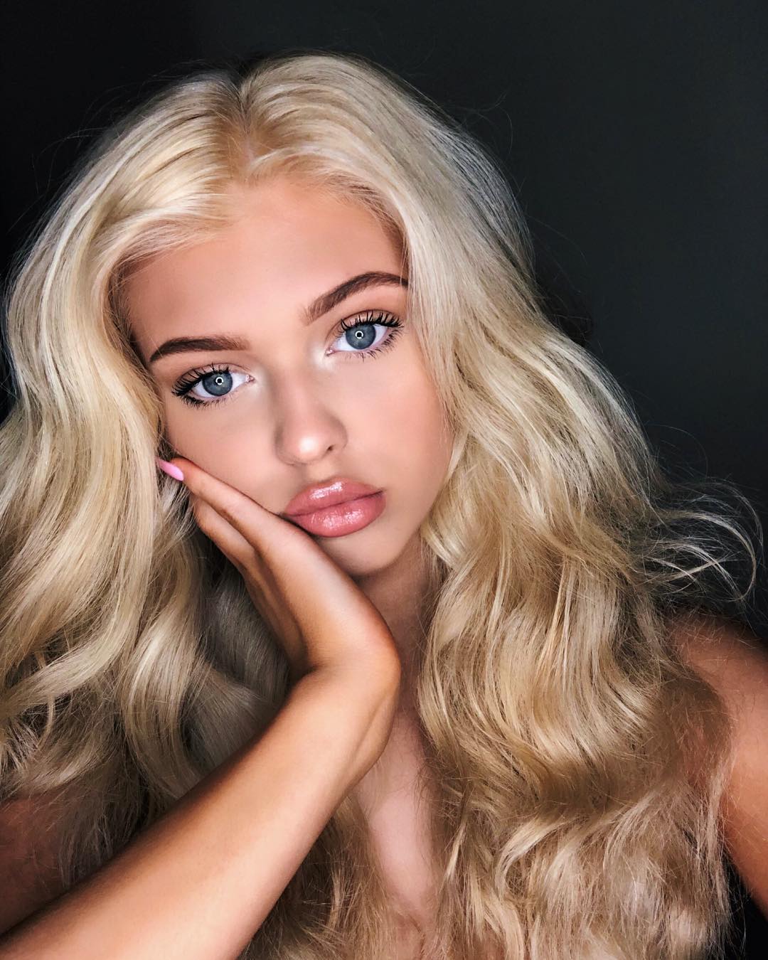 a portrait of a young woman with blonde wavy hair and blue eyes. she is posing with her hand on her face, and she is wearing makeup. her lips are glossy. the background is dark.