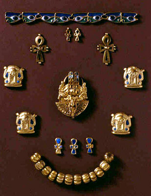 the image showcases a collection of ancient egyptian jewelry pieces arranged against a dark redbrown background. the artifacts, primarily made of gold and decorated with blue enamel, include a pectoral featuring a winged scarab beetle, various amulets shaped like the ankh symbol, protective deities, and a decorative bracelet and necklace with repeating gold beads. the arrangement suggests a museum display of these precious and historically significant artifacts.