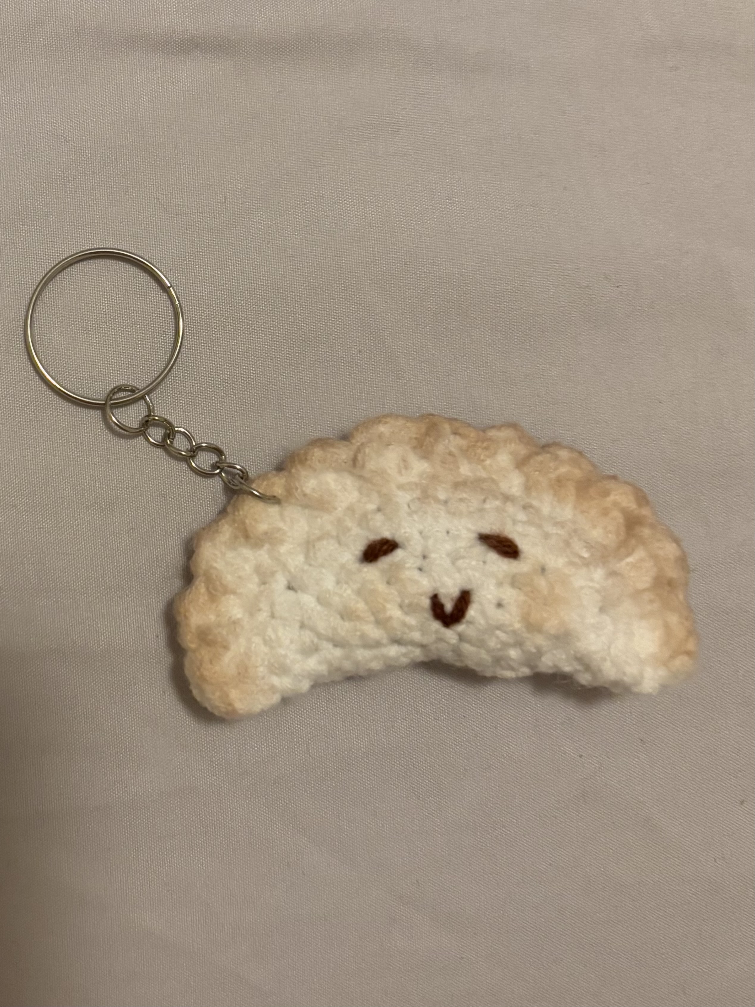 a closeup shot features a handmade crochet dumpling keychain lying on a flat, lightcolored surface. the dumpling is crafted from a soft, textured yarn in a pale offwhite color, with subtle hints of tan or peach around the edges to give it a realistic, cooked appearance. it's shaped like a crescent, with a crimped or scalloped edge mimicking the folded dough of a real dumpling. the dumpling has a simple, charming face embroidered onto it, consisting of two small, dark brown eyes and a downwardpointing "v" shape for a smiling mouth. attached to the top of the dumpling is a silver keychain, which includes a circular key ring connected to the dumpling by a short chain. the background is a plain, slightly textured surface, possibly a bedsheet or a fabric backdrop, which provides a neutral setting that allows the keychain to stand out.
