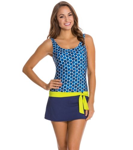 a woman is wearing a blue and white polka dot skirted onepiece swimsuit with a green sash. she is posing for the camera with her arms at her sides. she has long, dark hair. the background is white.