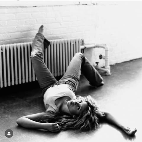 a black and white image features a woman lying down on a floor next to a radiator. she is wearing jeans and a tank top, and her hair is wavy. one leg is extended up against the radiator. she is looking at the camera. the background includes a brick wall and pipes. the scene is lit with a combination of light and shadow.
