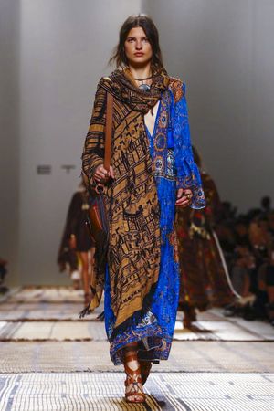 a model walks down a runway wearing a long, blue and brown patterned dress with a matching shawl and a brown leather handbag. her look is bohemian and hippiechic. the background is blurred but shows other models and a crowd of people. the setting appears to be a fashion show.