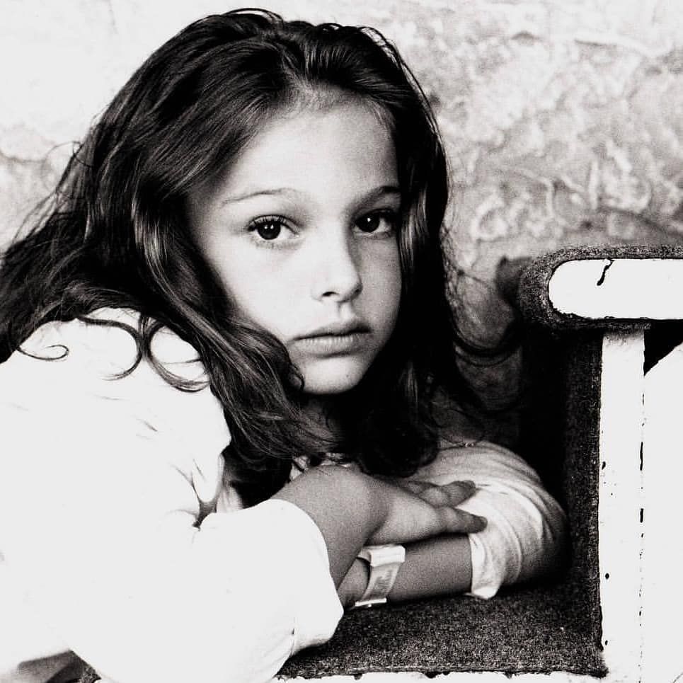 a black and white portrait of a young girl, possibly natalie portman, looking at the camera with a serious expression. she has dark hair and is wearing a lightcolored shirt. her arms are crossed and she's wearing a watch. she's leaning against a chair. the background is slightly blurred.