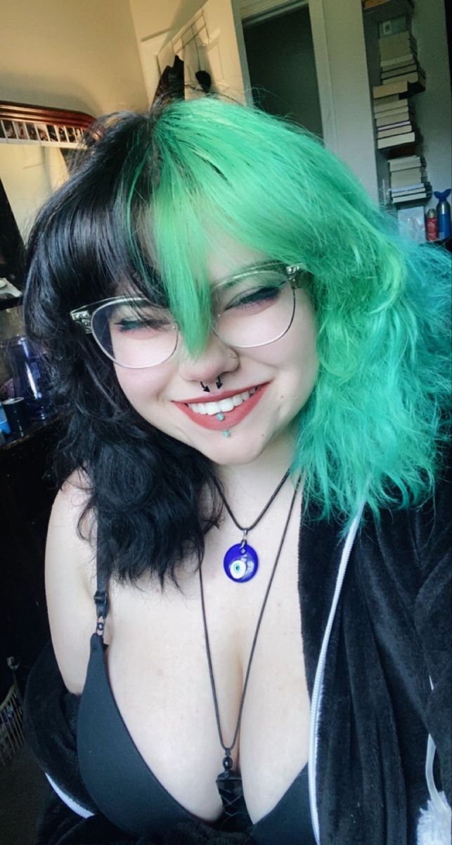 a woman with halfdyed hair (green and black), wearing glasses, a bra, and necklaces, is smiling. she has piercings on her nose and lip.