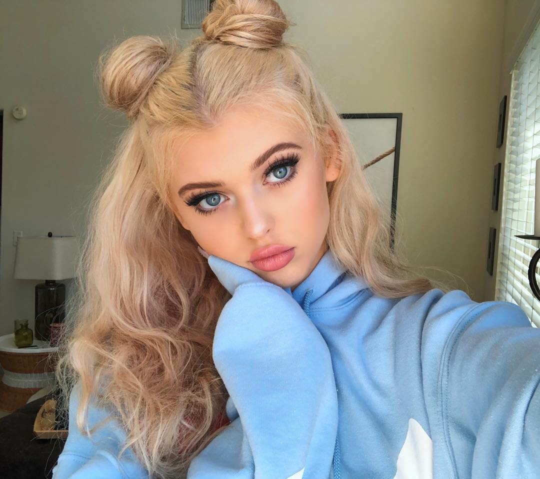a selfie of a blonde woman with her hair in two buns on top of her head and long, wavy hair framing her face. she is wearing a light blue hoodie and has her hand resting on her face. she has striking blue eyes, long eyelashes, and pink lipstick. the background shows an indoor setting with a lamp, table, plant, picture frame, and blinds.
