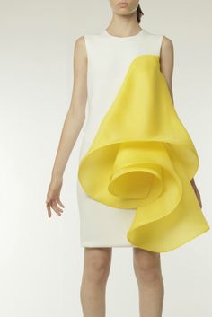 a woman is wearing a white sleeveless dress with a large yellow ruffle that is shaped like a rose. the dress is fitted and the ruffle is very full and dramatic. the model is posing in a studio with a white background. the lighting is soft and flattering.  the dress is modern and sophisticated, and the woman looks elegant and stylish.