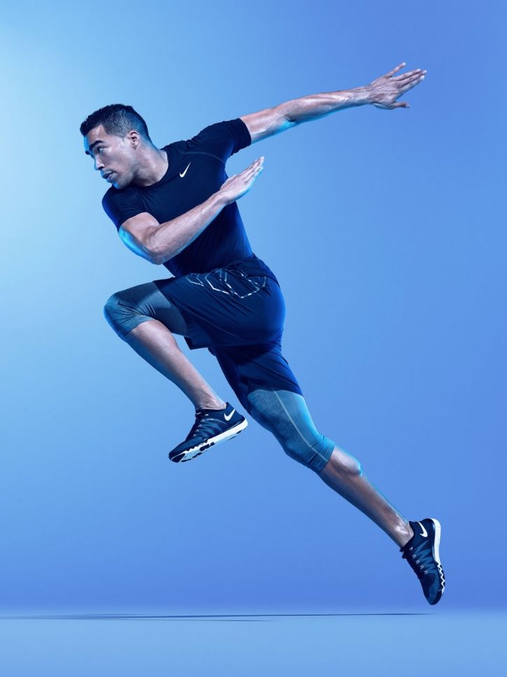 a man in a black nike shirt, black shorts, and black nike shoes is midair while jumping. the image is in a blue studio setting.