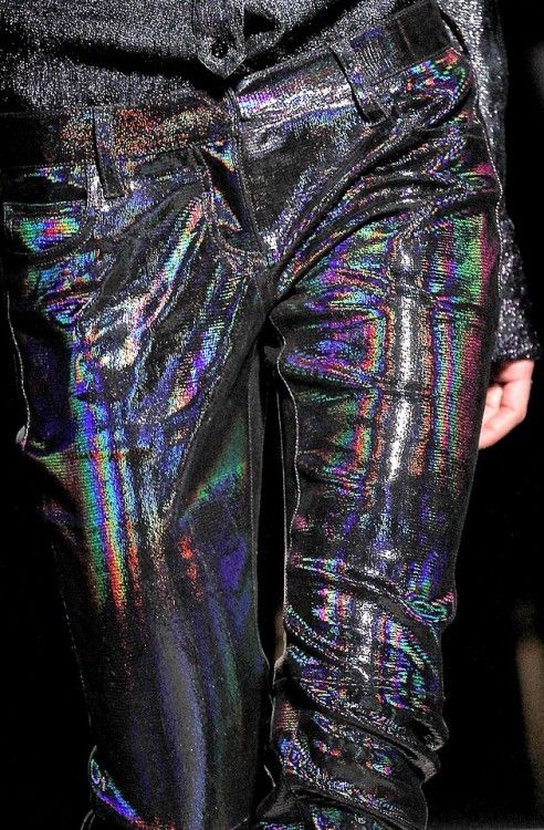 a close up of a model wearing shiny, iridescent black jeans on a runway. the jeans are made of a material that reflects light in a rainbow of colors. they are tightfitting and have a sleek, modern look.