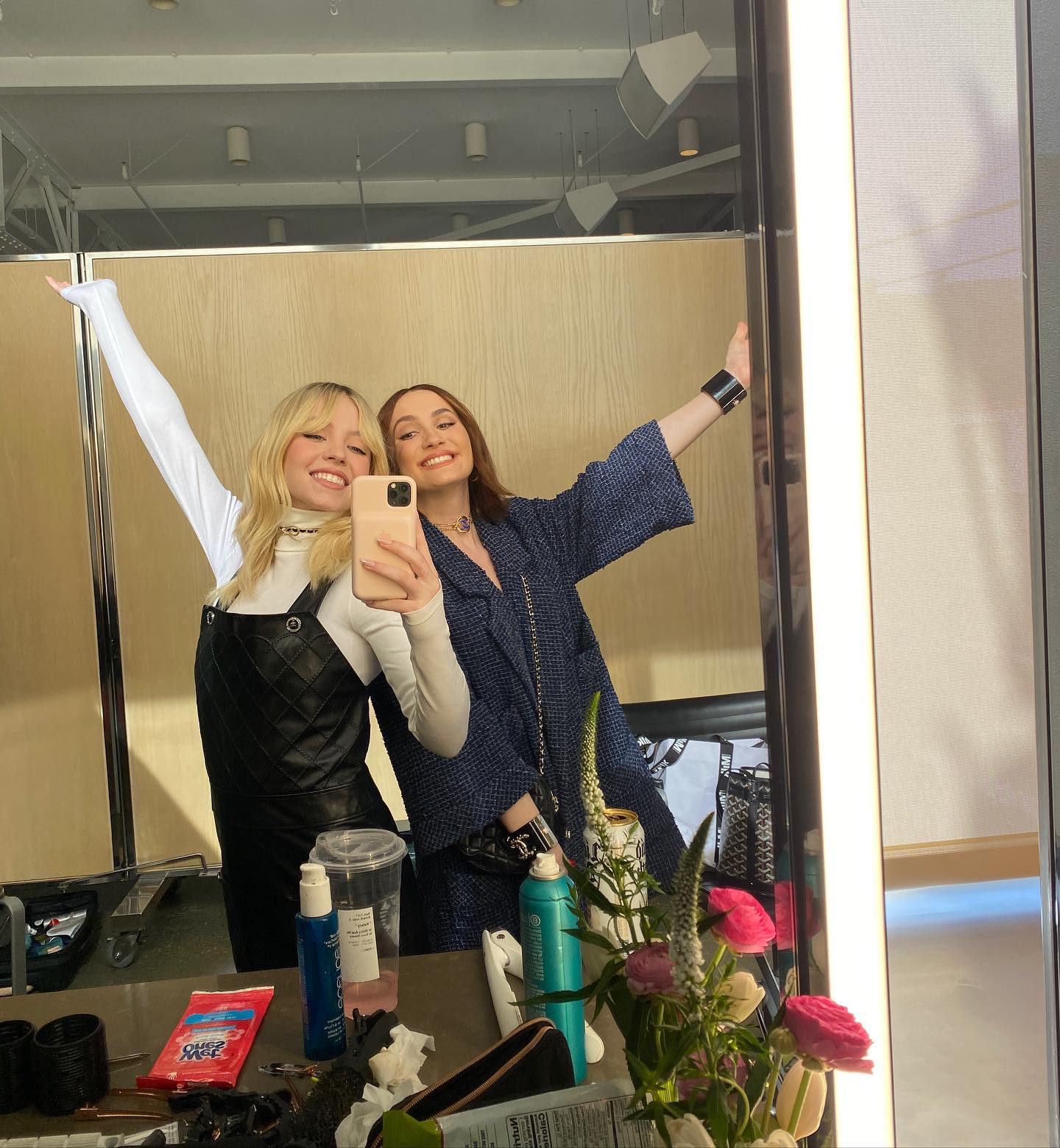 a selfie taken in a mirror shows two young women backstage, possibly in a dressing room. one girl has blonde hair and is wearing a black quilted dress and white longsleeved shirt.  the other girl has darker hair and is wearing a blue tweedstyle jacket. they are smiling and appear happy.  various hair products and makeup items are on the vanity in front of them, along with a bouquet of pink and white flowers. the setting suggests a professional environment like a photoshoot or a television show. the overall mood is light and cheerful.