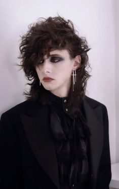 a young woman with long, dark, curly hair is looking down at something in a dark and moody portrait. she is wearing all black clothing, including a black blazer, a black shirt with frills, and black earrings.  she has a choker around her neck.  she is posing against a white background. the style is gothic and alternative.