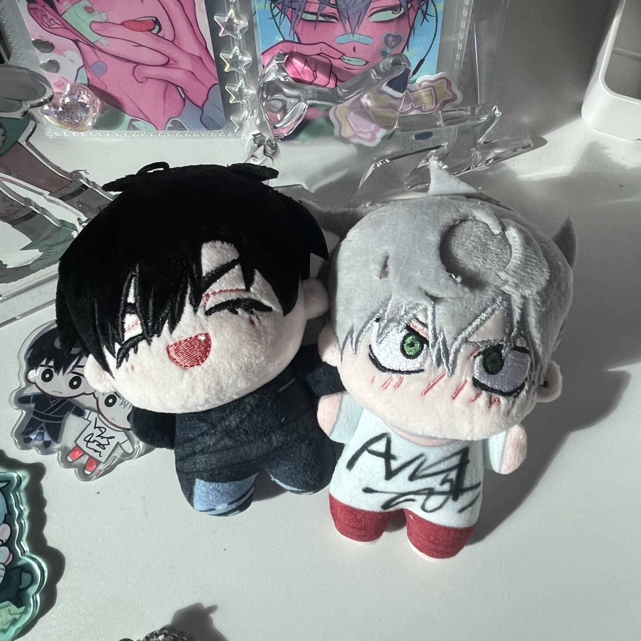 two anime plush toys, one a chibi character with black hair, the other with white hair, sitting on a table. they are both smiling and looking to the right. the character with black hair is wearing a black jacket with blue jeans and the white hair character is wearing a white shirt with red pants. there are other anime toys in the background and a white and gray surface the plush toys are on.