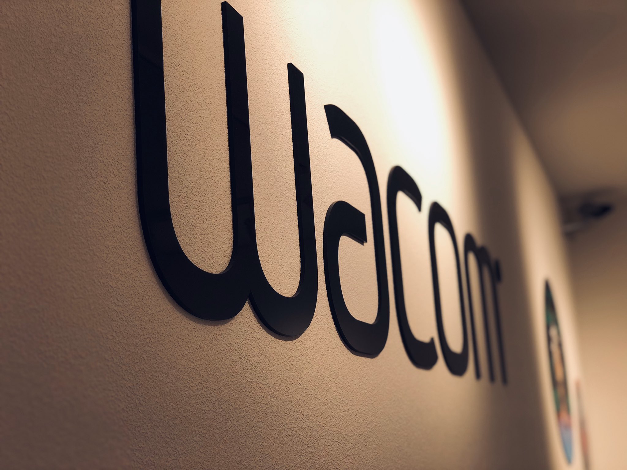 a closeup shot of a black wacom logo mounted on a textured beige wall. the letters cast subtle shadows on the wall, indicating artificial lighting.  the perspective is at eyelevel, drawing focus to the brand name and its association with creativity and digital art.