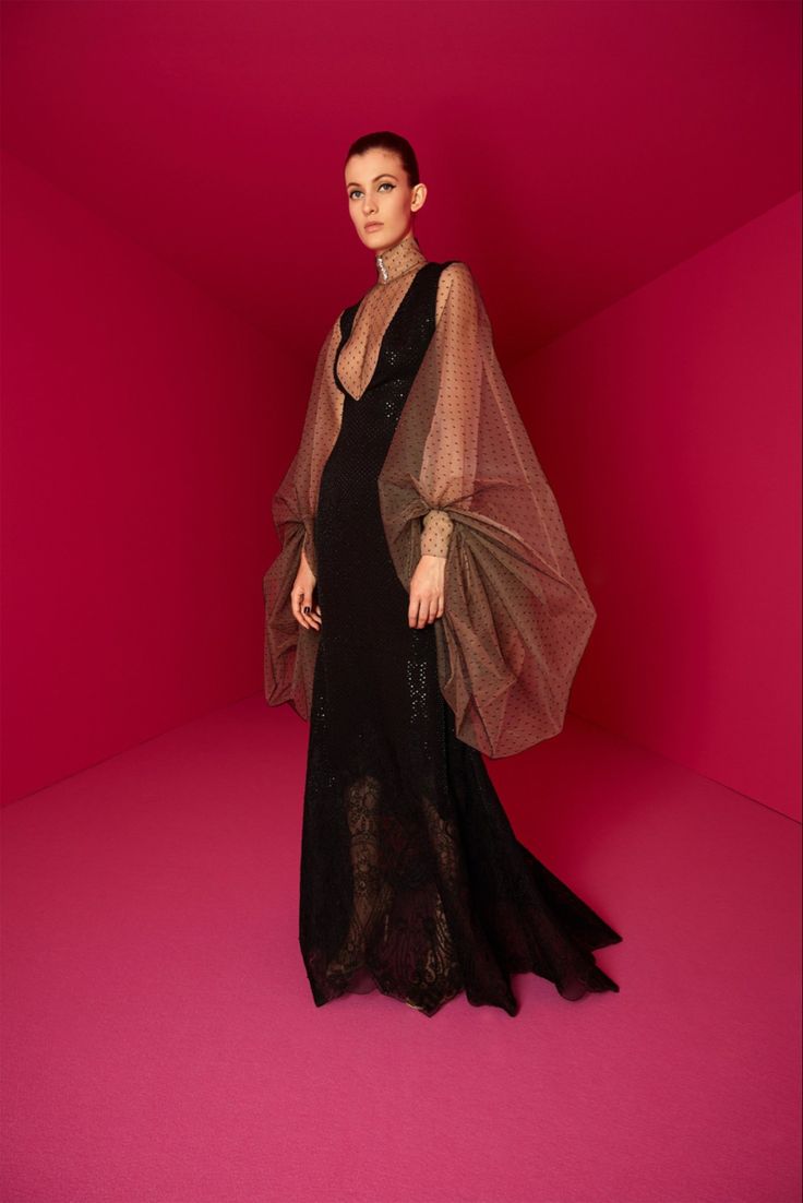 a woman is wearing a black gown with sheer, polkadotted sleeves in a red room. she is looking slightly to the left of the camera. the dress has a plunging neckline and a fitted bodice, flaring out at the bottom. the sleeves are very large and billowy, gathered at the wrists. the background is a solid red, with the walls and floor meeting at sharp angles.