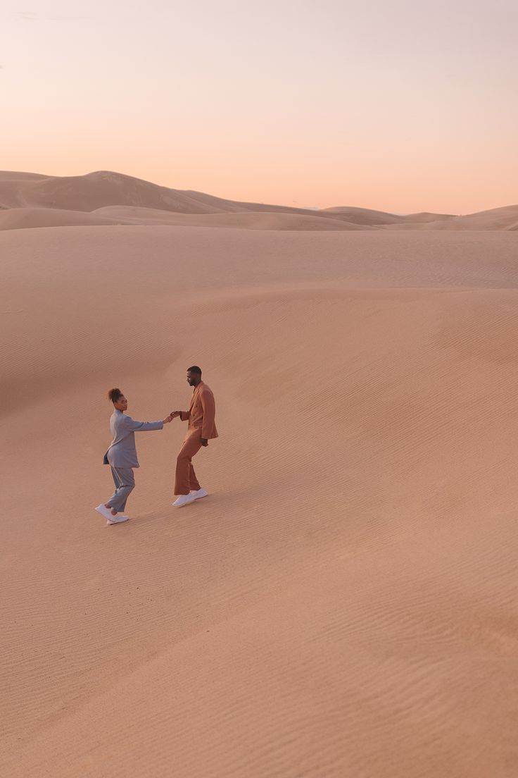 a couple in stylish outfits are walking hand in hand on a sand dune in the desert. the man is wearing a brown suit and the woman is wearing a blue suit. the sun is setting in the background, creating a golden hour glow. the sand dunes stretch out as far as the eye can see. they look happy and in love, and the image captures a sense of adventure and romance.