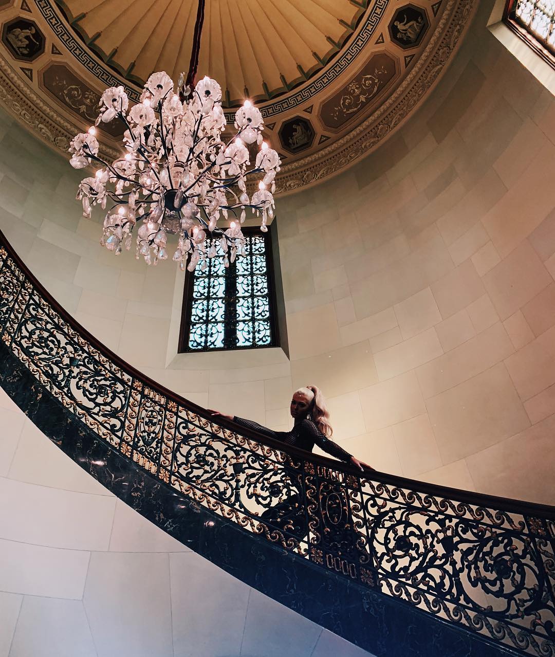 a lowangle shot captures a woman standing on an ornate, marble staircase inside a grand building. above her hangs a large, crystal chandelier, with a decorative dome ceiling in the background. a gothicstyle window is also visible near the chandelier.