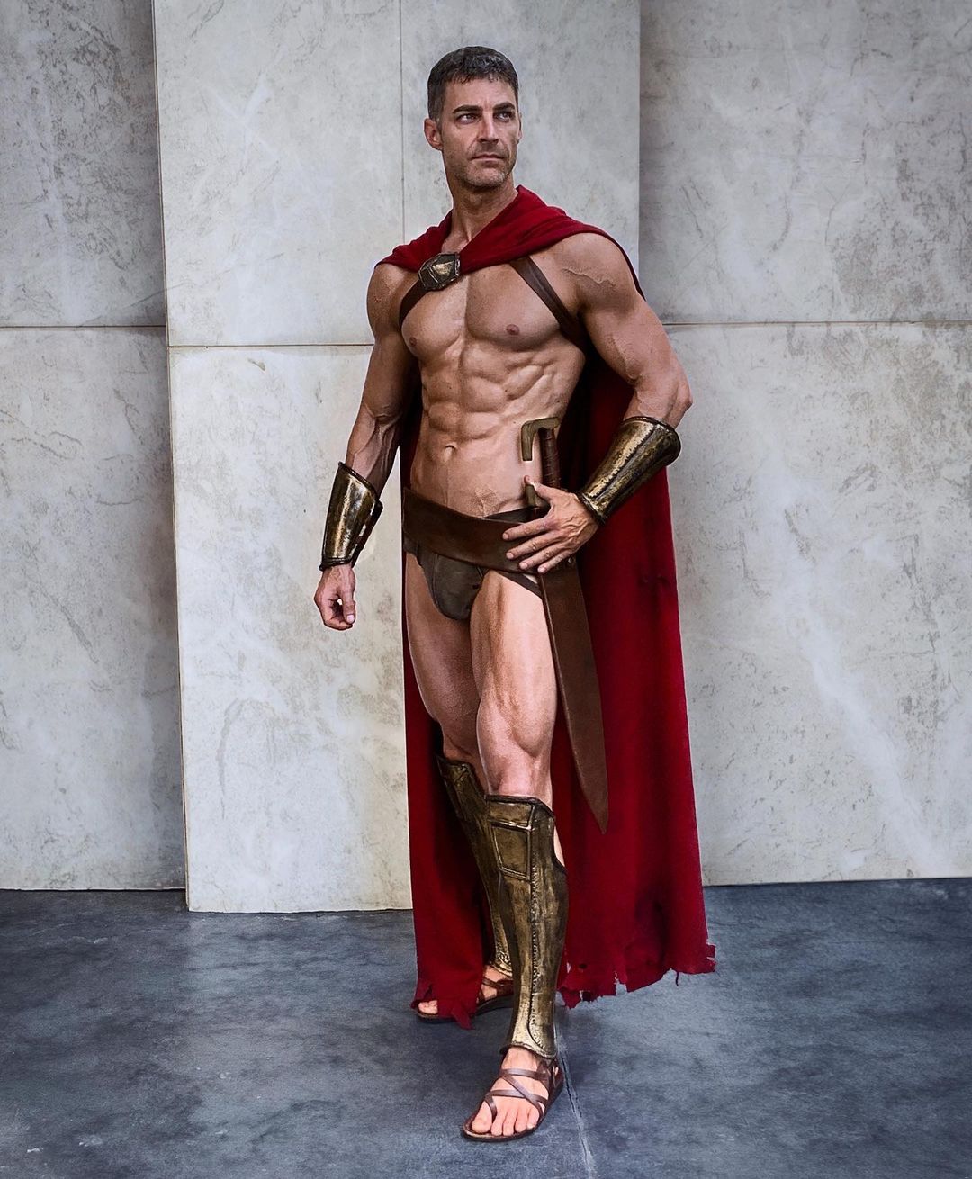 the image shows a muscular man dressed in a meticulously detailed spartan warrior costume.  he is wearing a red cape, gold greaves (leg armor), and a brown loincloth.  his physique is extremely defined, suggesting bodybuilding or fitness modeling. the setting appears to be a studio with a textured, light gray background. the man's pose is confident and suggestive of a warrior's stance. the costume is highly realistic, suggesting a high level of effort in its creation, possibly for a cosplay event or photoshoot.  the overall style strongly evokes the imagery of the movie "300."