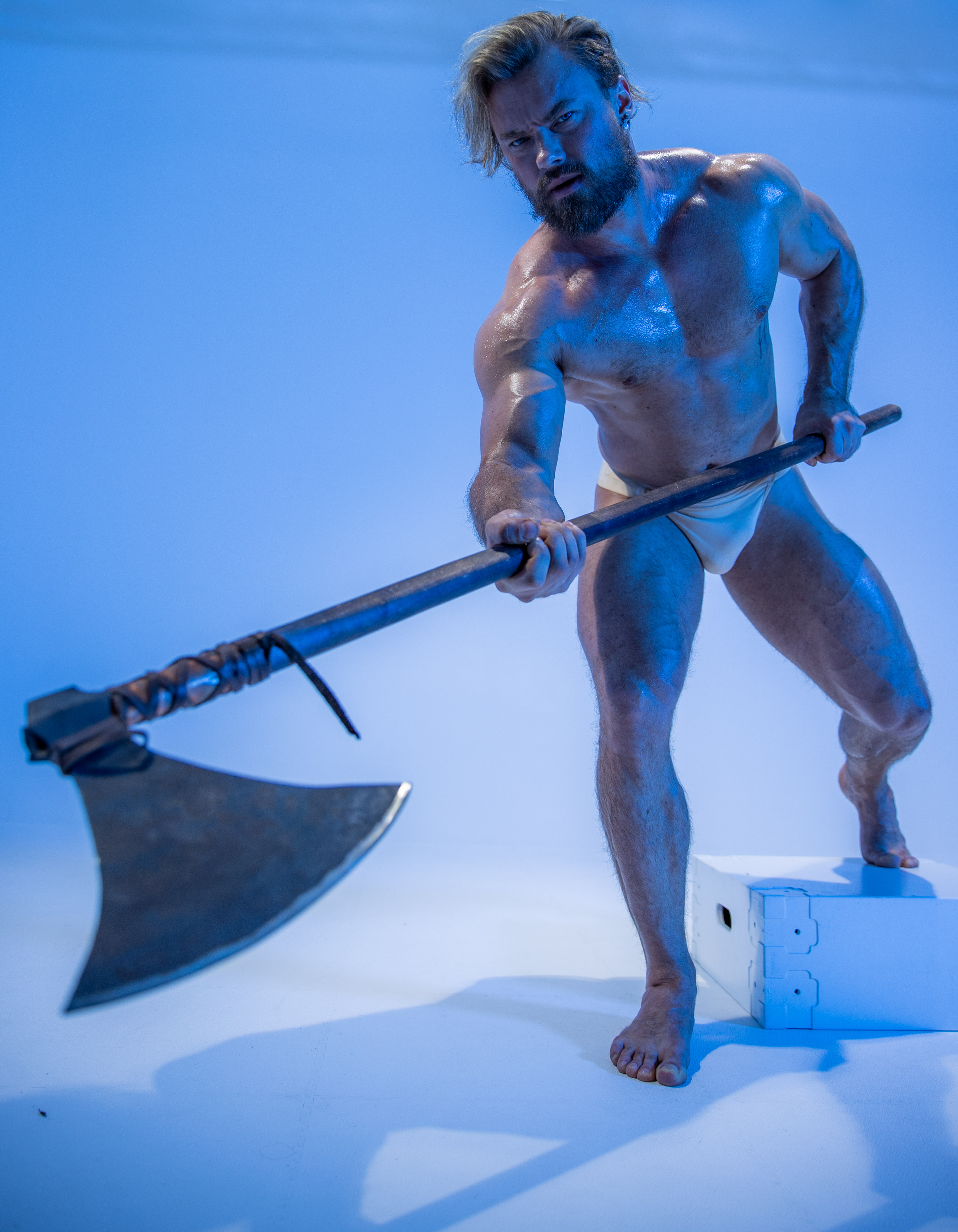 a muscular man with long blond hair and beard, wearing only a loincloth, stands in an aggressive pose against a bright blue background. he holds a large, doubleheaded axe in his hands, raised as if ready to strike. the lighting is bright and artificial, giving the image a dramatic and powerful feel. he is standing on the balls of his feet with one foot on a white box.